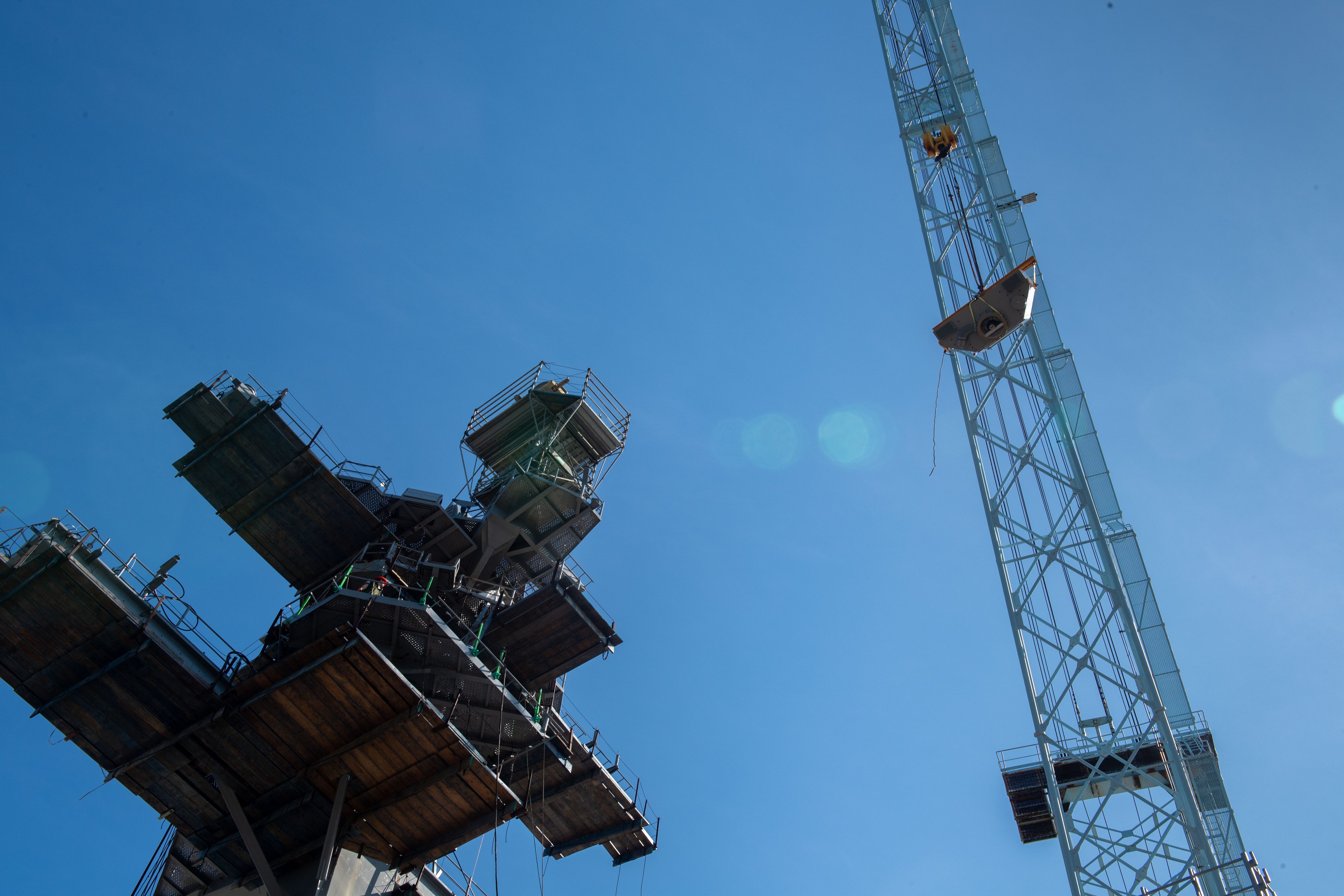 A view from under the tower of an aircraft carrier and a crane. The sky in the background is clear blue.