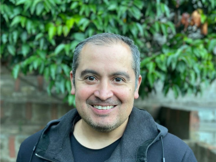 A man smiling outdoors in front of green foliage, wearing a black zip-up jacket over a dark shirt.