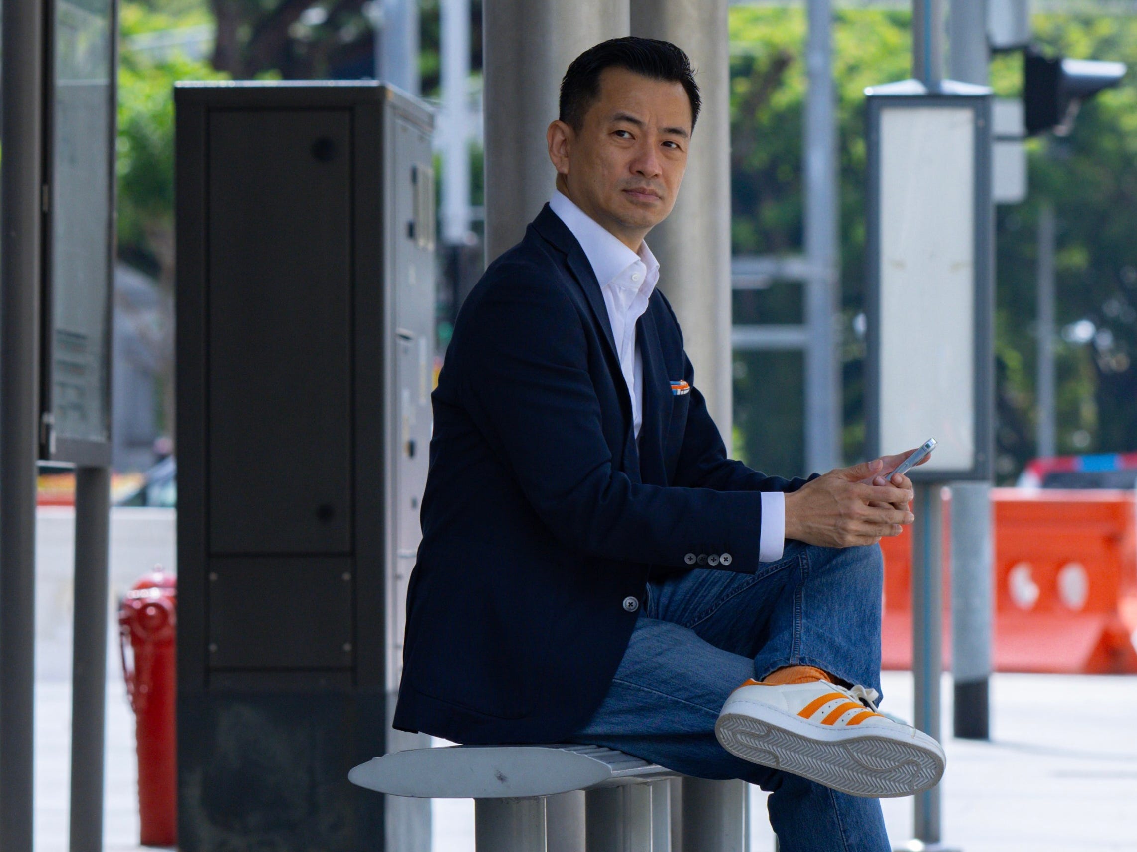 Eric Sim is wearing a blue blazer and sitting at a bus stop.