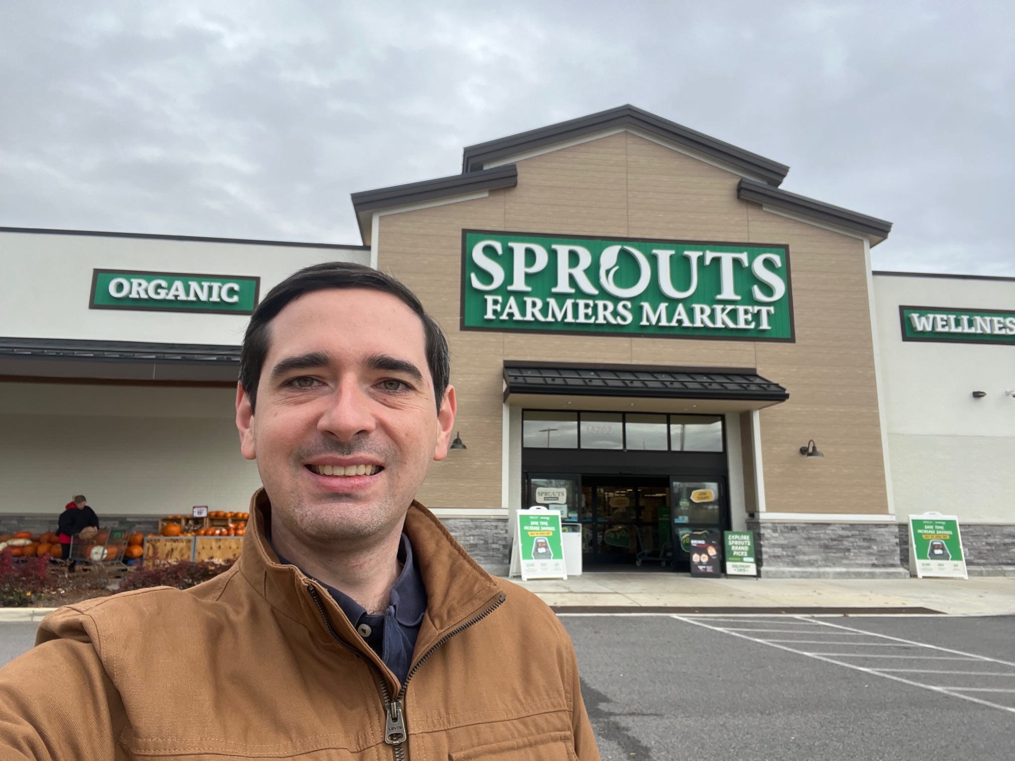 Business Insider reporter Alex Bitter wears a khaki-colored coat while standing in front of the entrance to a Sprouts Farmers Market Store in Maryland.