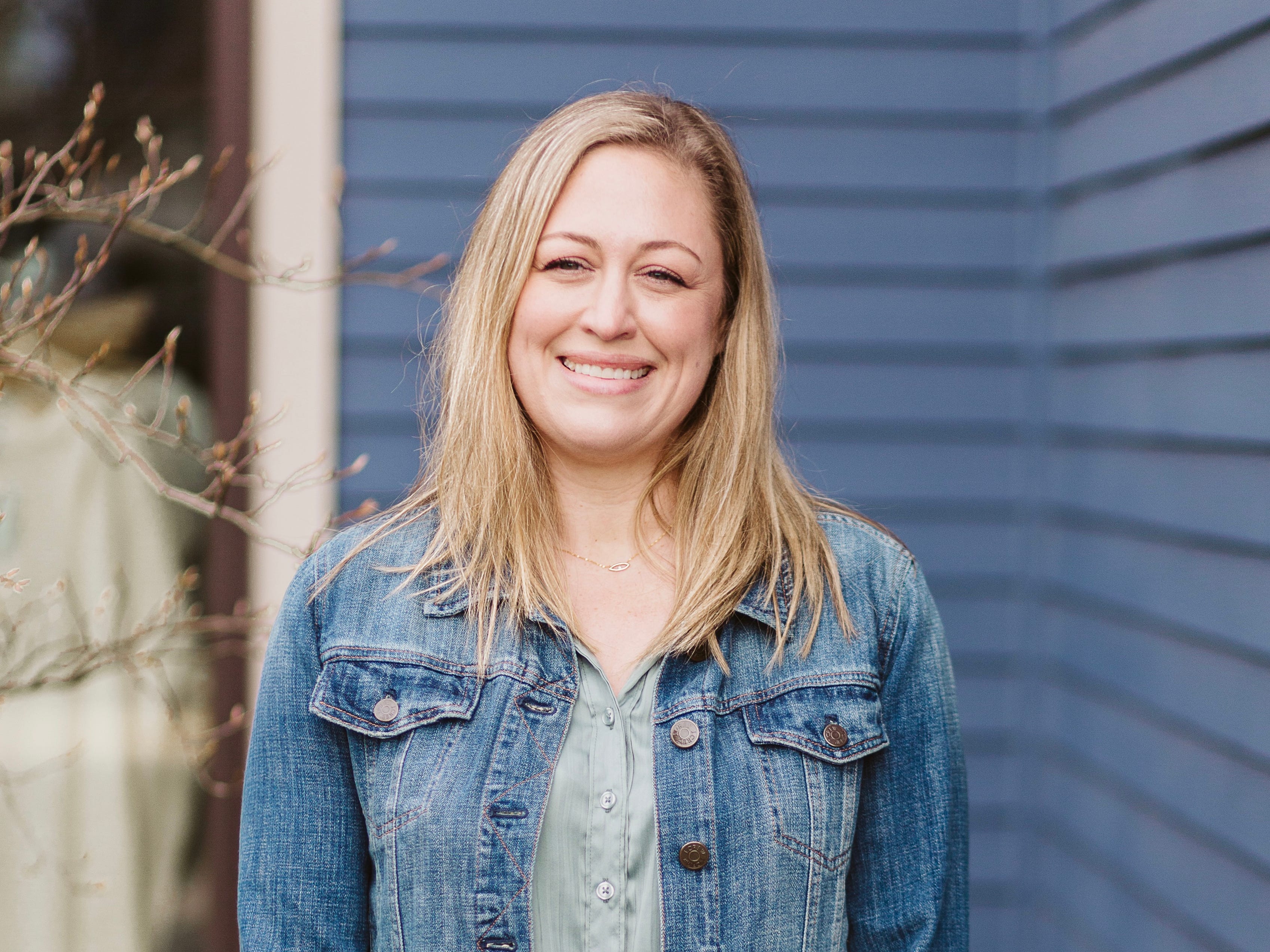 a woman poses in front of a blue house in a denim jacket