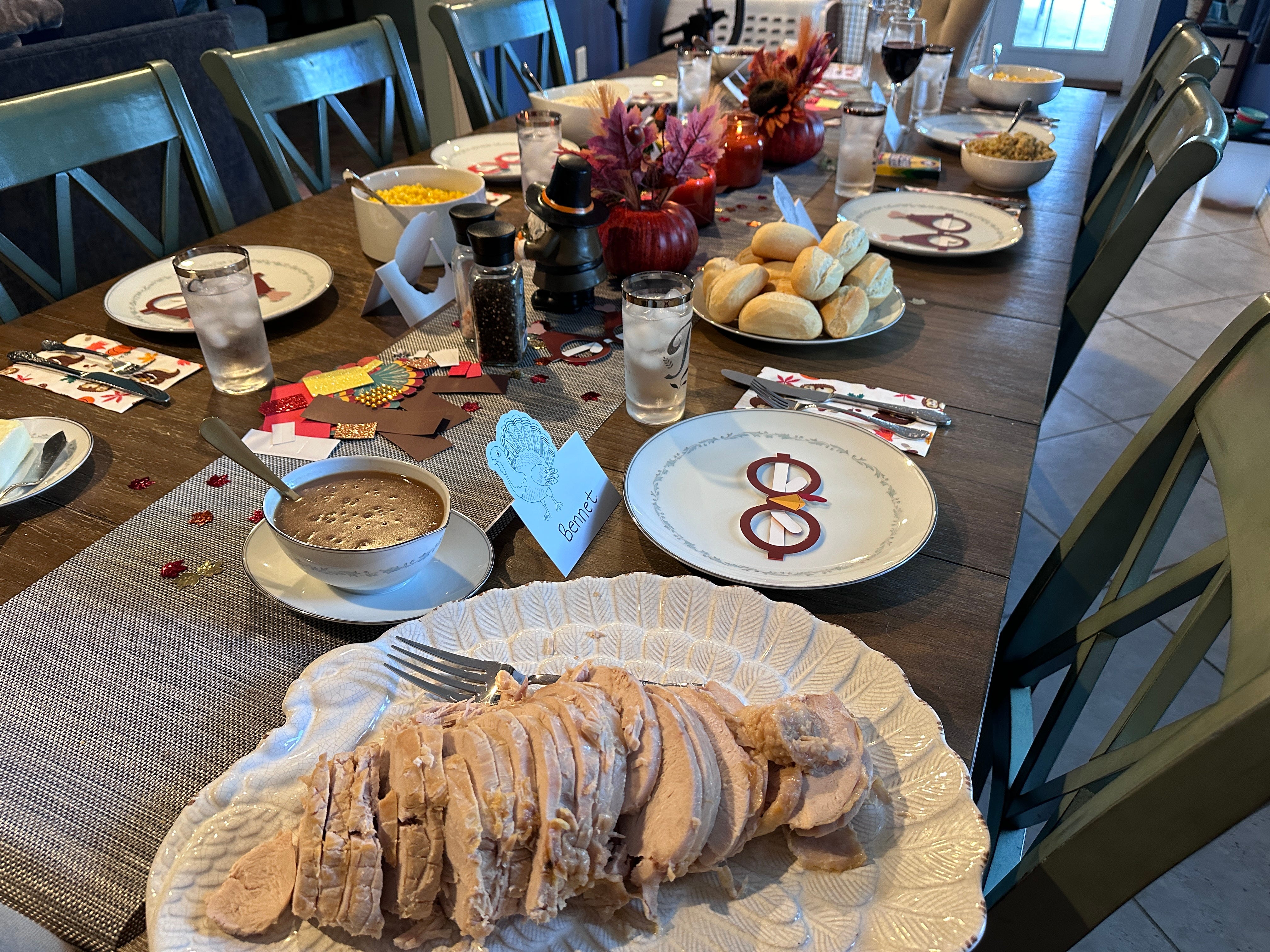 Sliced turkey, a plate of rolls, a bowl of gravy, and other Thanksgiving foods on a table set for a Thanksgiving dinner