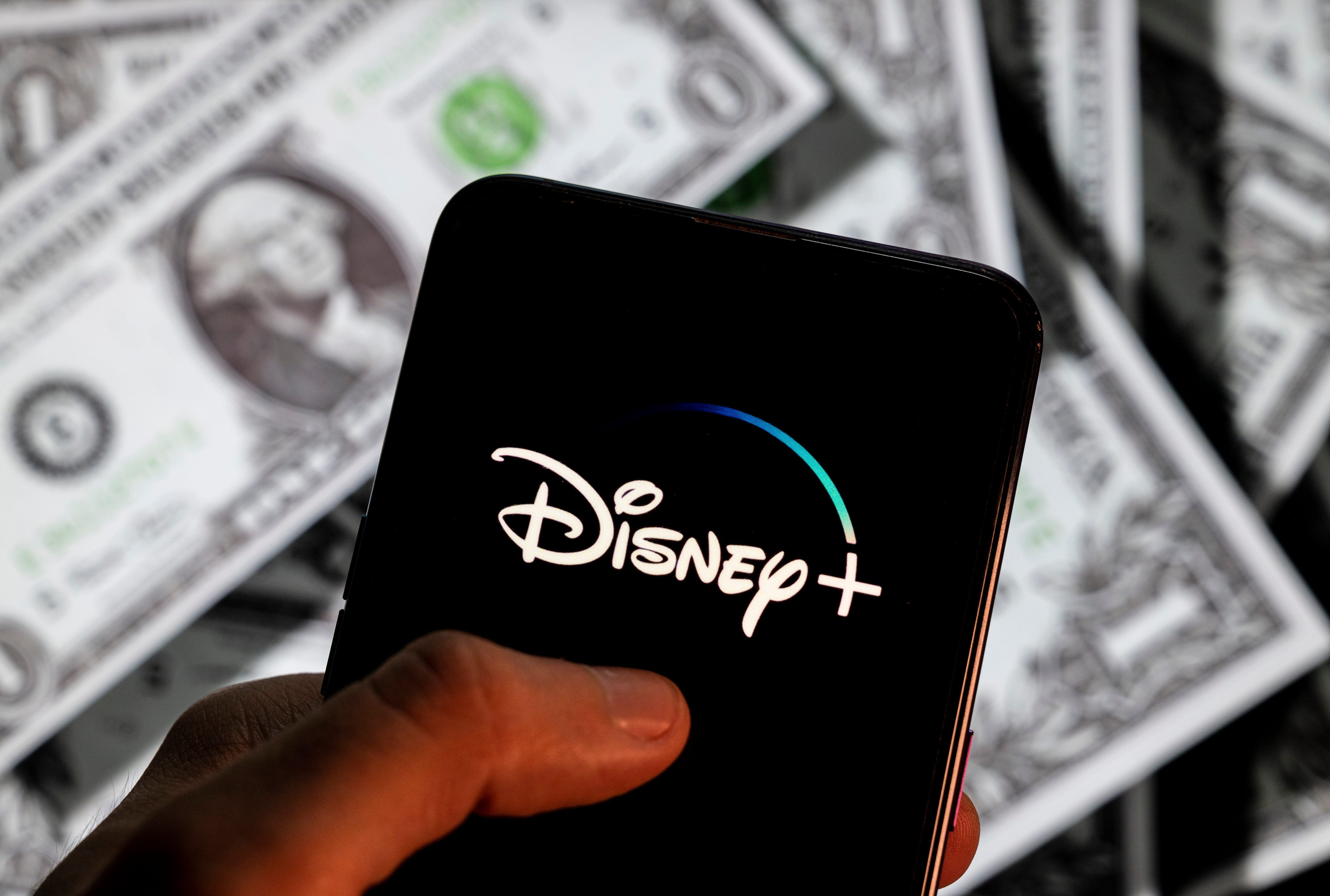 The Disney Plus logo is displayed on a smartphone screen in someone's hand with several $1 bills blurred in the background.