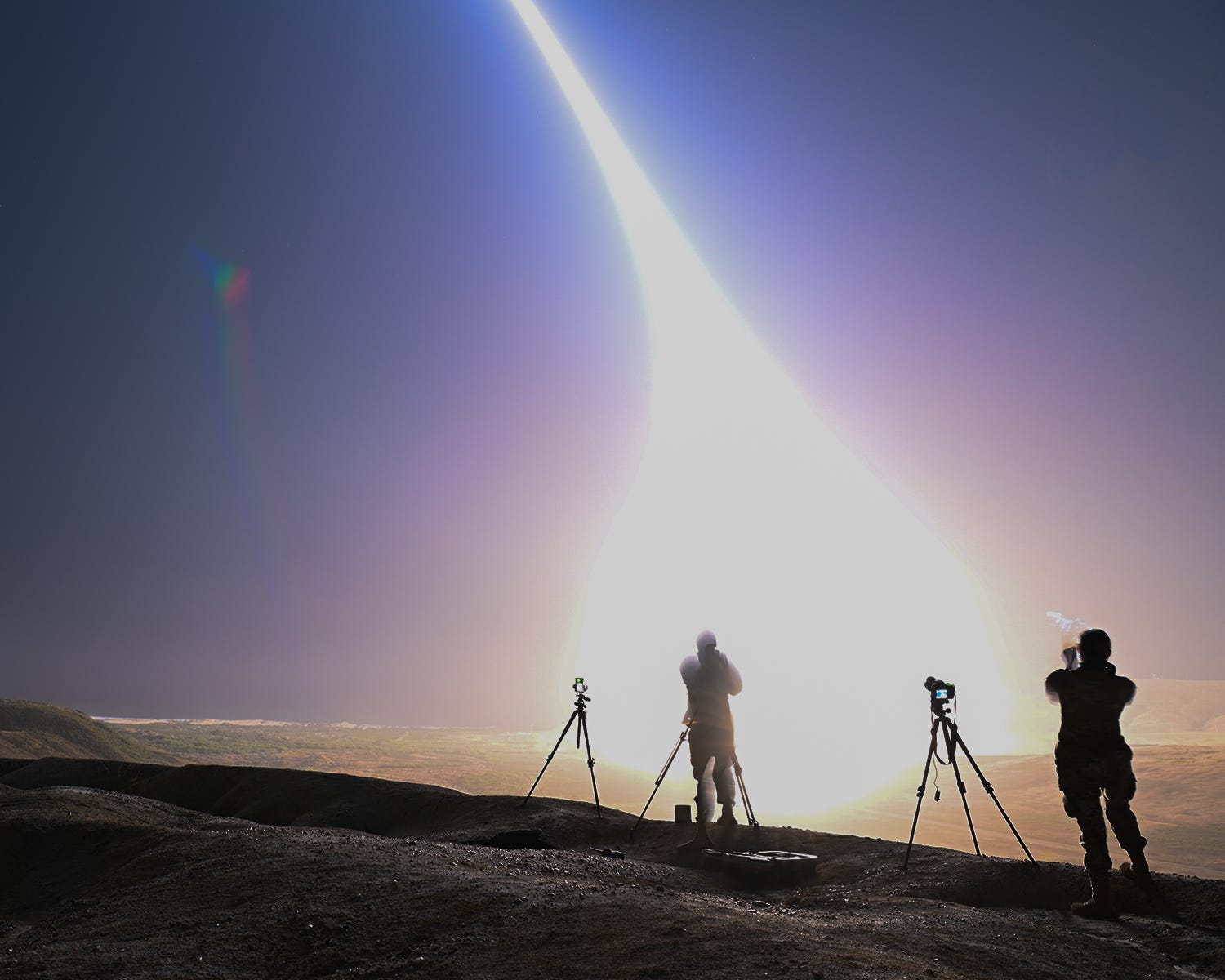 Two men stand on a barren field as an intercontinental ballistic missile launches into the sky.