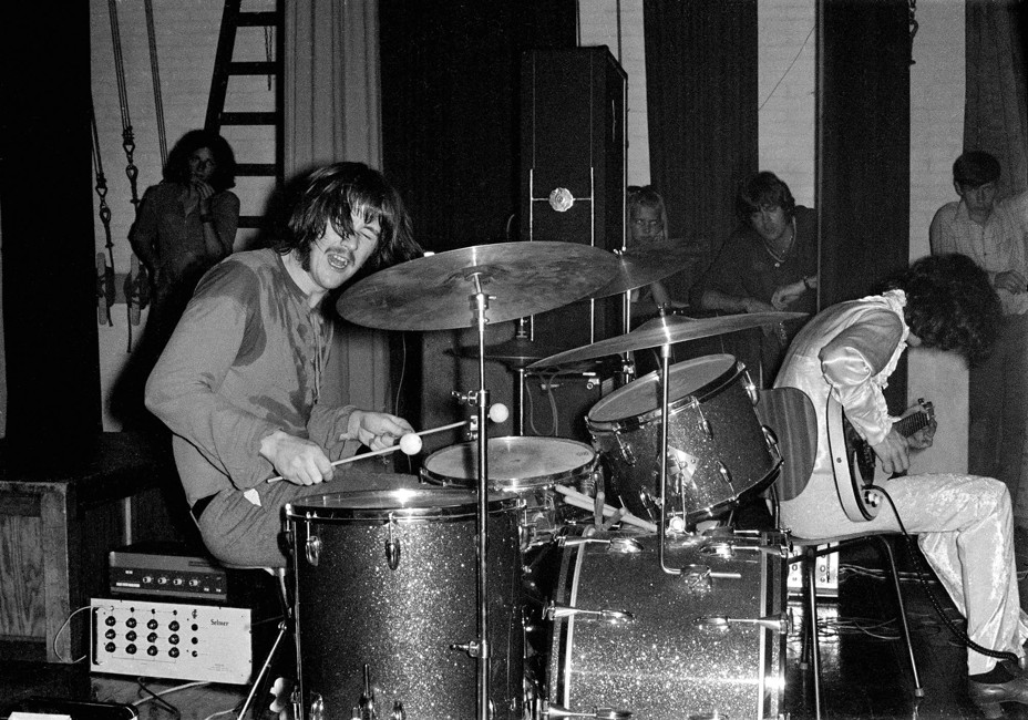 black-and-white photo of man sweating through his shirt playing drums next to man in white bent over his electric guitar