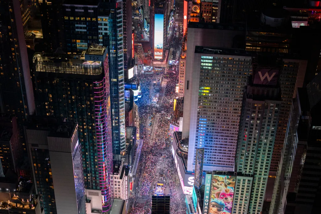 Aerial photo of Times Square at night during New Year's Eve, with large crowds filling the streets surrounded by brightly lit skyscrapers and electronic billboards.