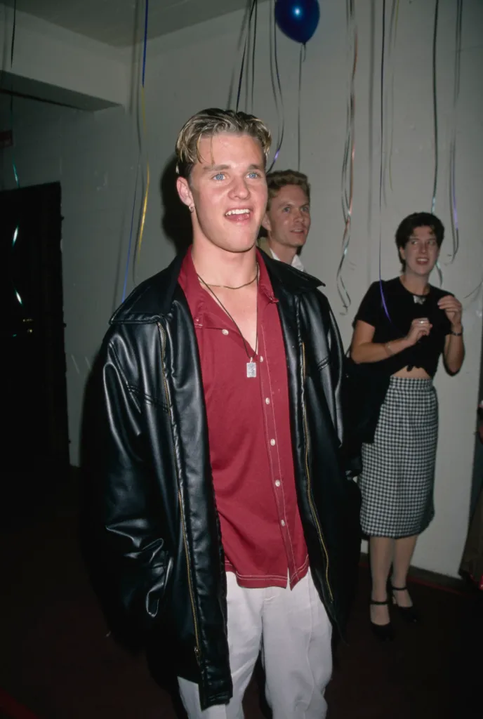 American actor Zachery Ty Bryan in a red shirt, black leather jacket, and white trousers.