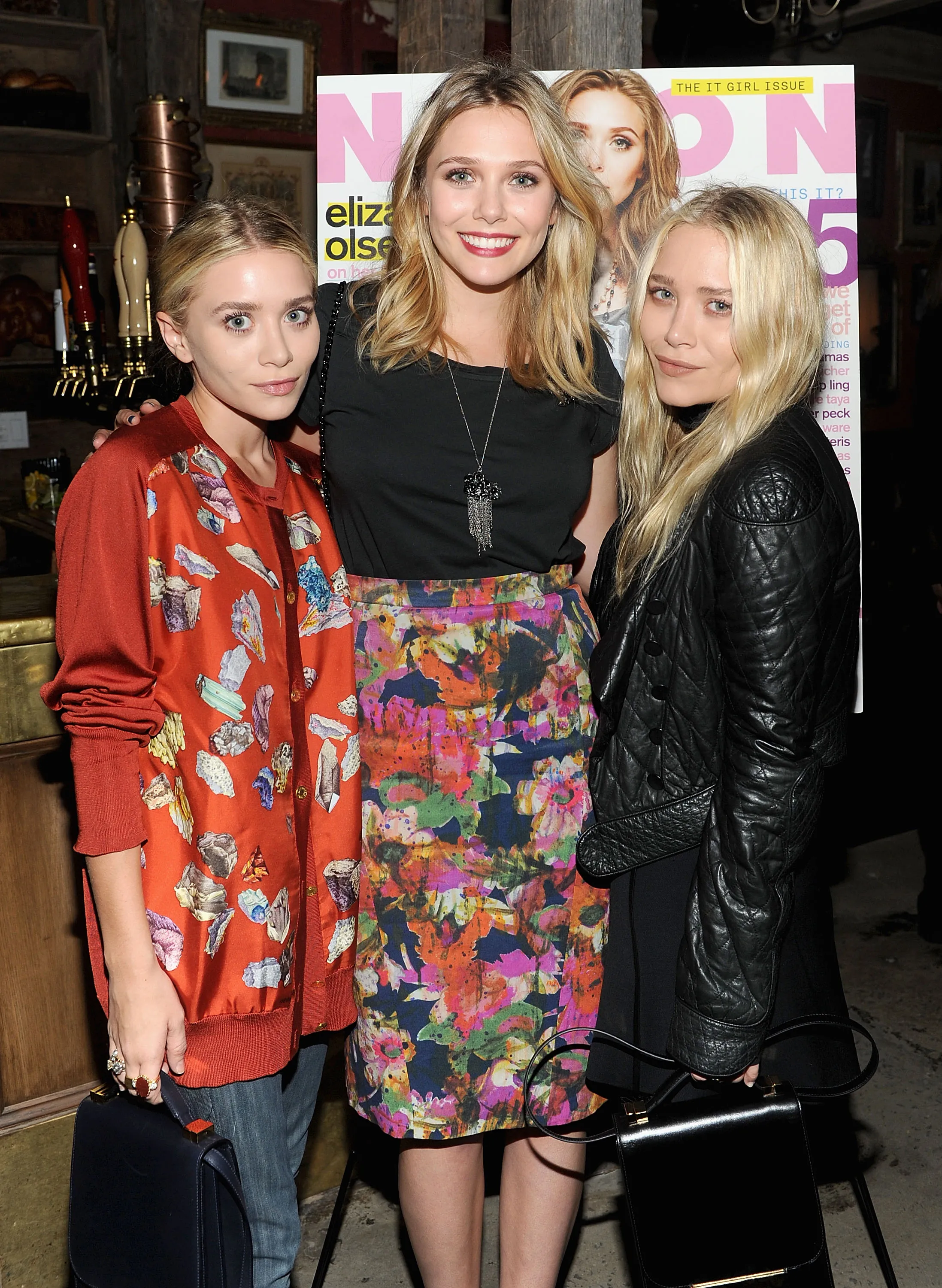 Ashley Olsen, Elizabeth Olsen, and Mary-Kate Olsen standing in front of a Nylon Magazine poster.