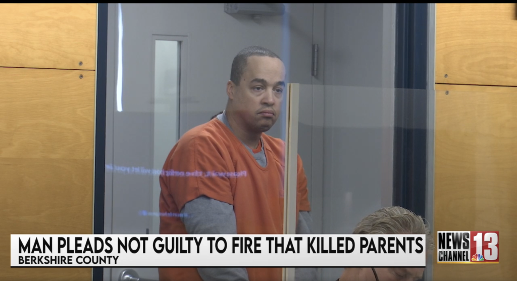 Darius Hazard in an orange jumpsuit behind a clear divider, with a news chyron at the bottom reading 