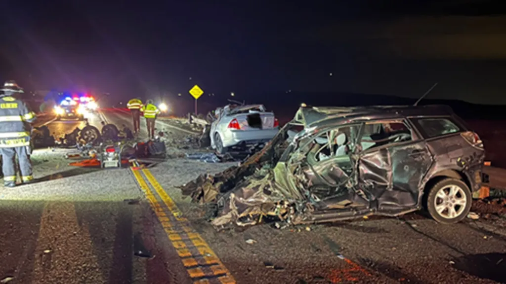 Nighttime photo of a highway accident scene with severely damaged vehicles and emergency personnel.