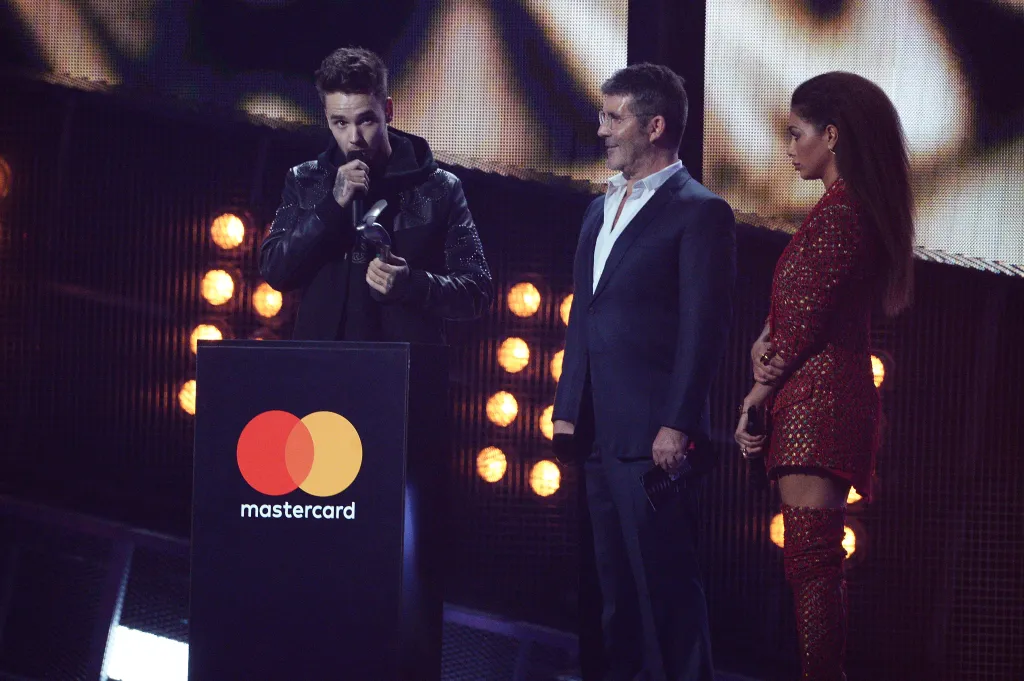 Liam Payne speaking at a podium with the Mastercard logo, next to Simon Cowell and Nicole Scherzinger, at The BRIT Awards 2017.
