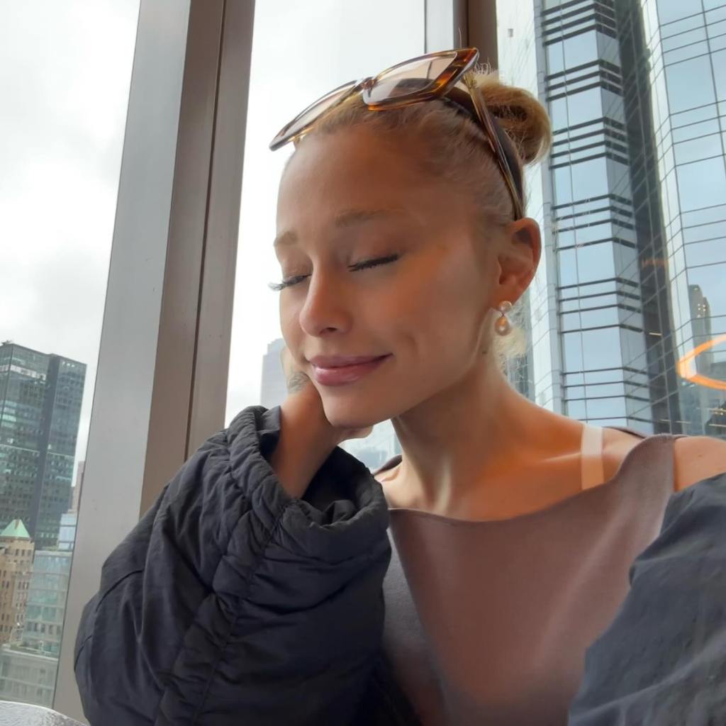 Ariana Grande with her eyes closed, wearing a dark jacket, sitting by a window overlooking city buildings.