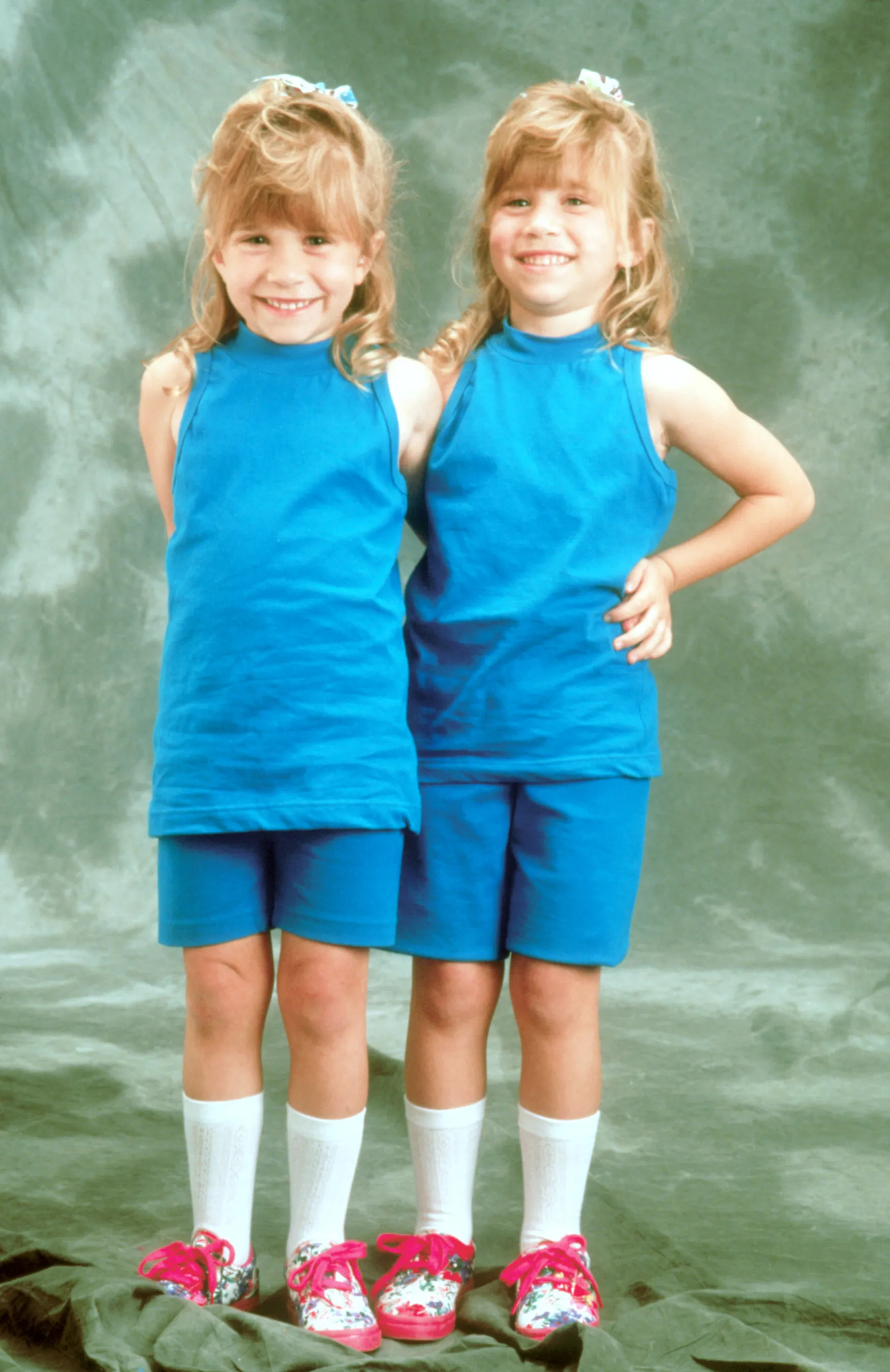 Mary-Kate and Ashley Olsen, as child actors, smiling while wearing matching blue outfits and shoes.