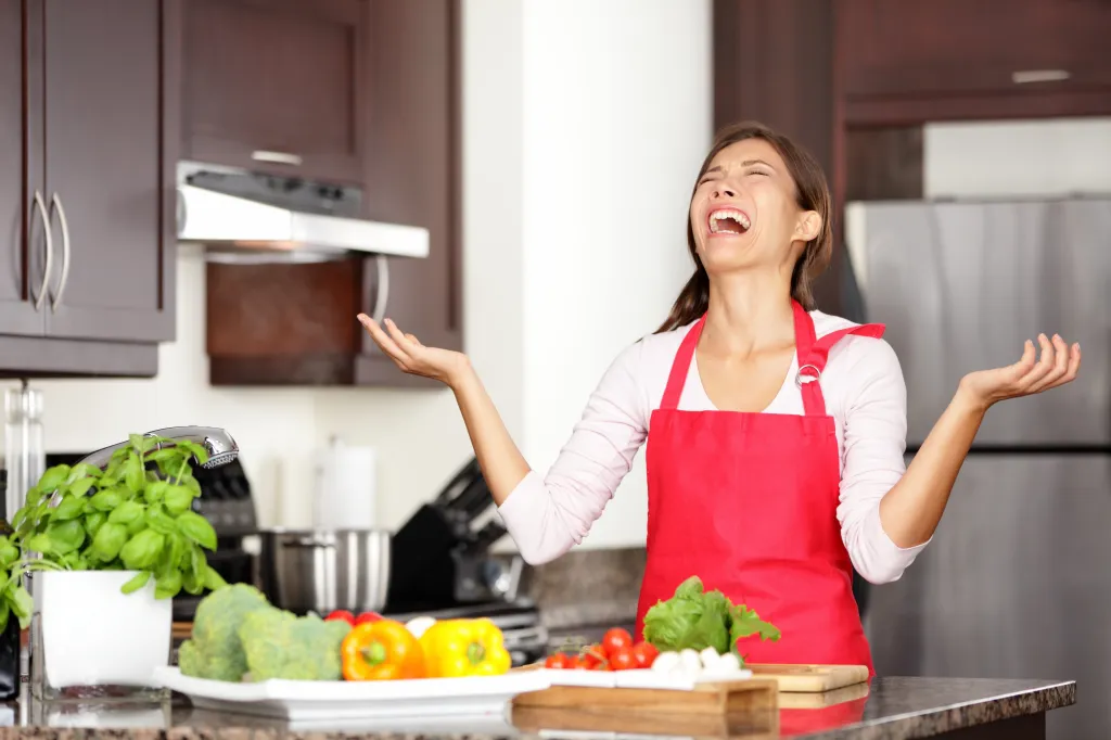 A frustrated woman in a red apron throws her hands up in the kitchen, surrounded by vegetables.