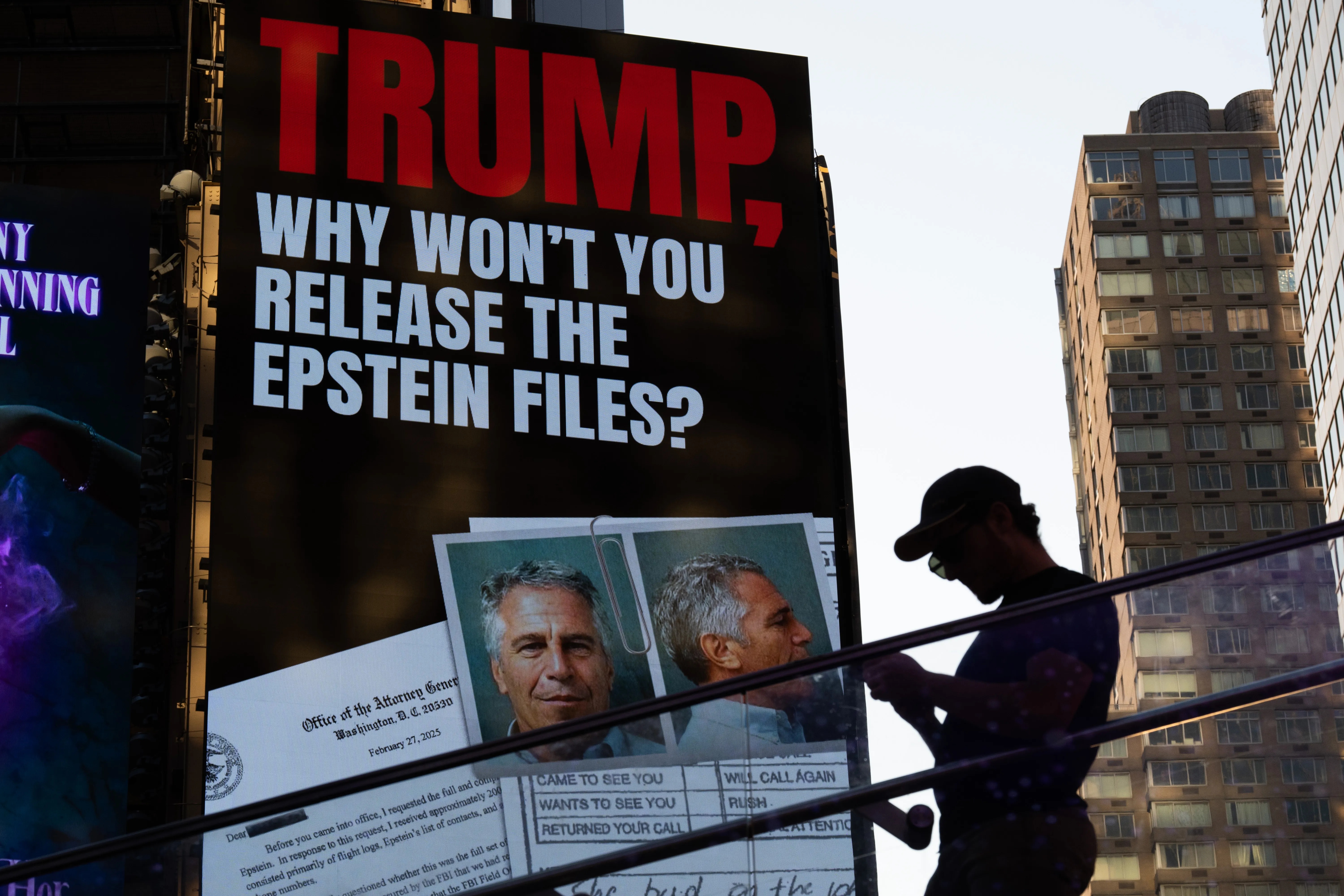 A large black billboard reads “Trump, why won’t you release the Epstein files?” while a man in a baseball cap stands in front of it.