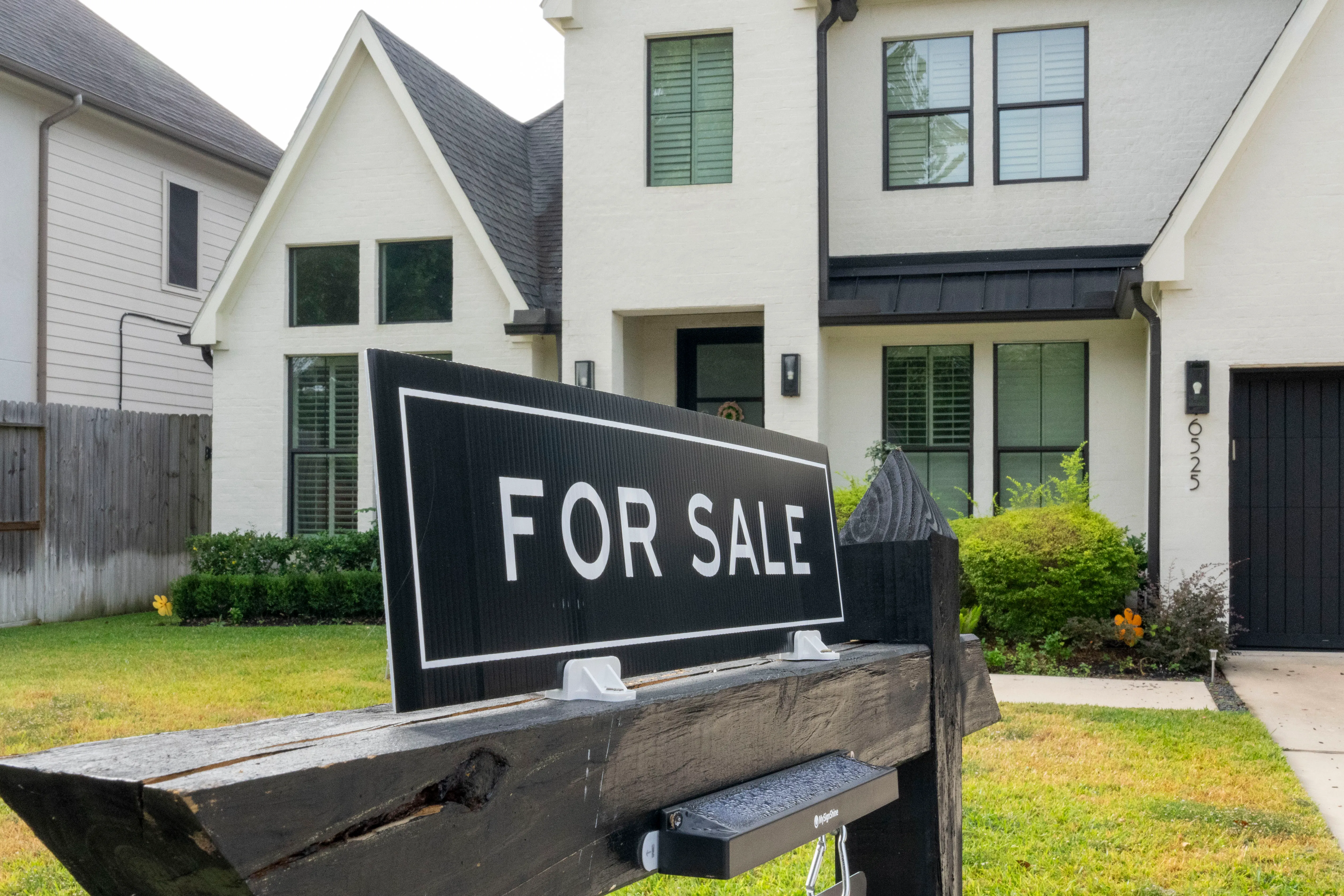 A black FOR SALE sign looms in from its position on the lawn of a large white suburban house.
