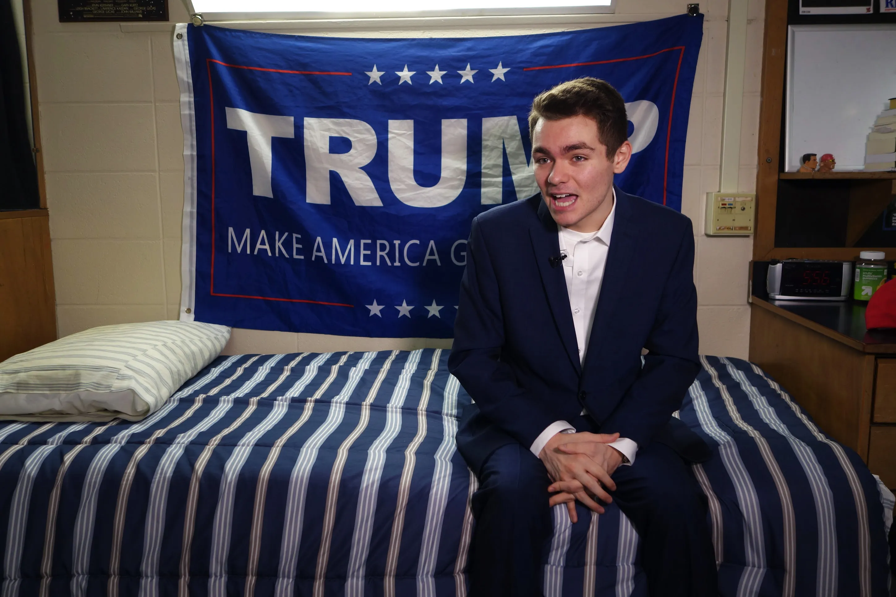 Nick Fuentes sitting on a bed and talking with a Trump MAGA banner hanging behind him