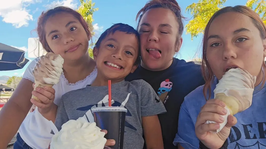 Four people, including young children, eating ice cream cones and drinking a beverage outdoors.