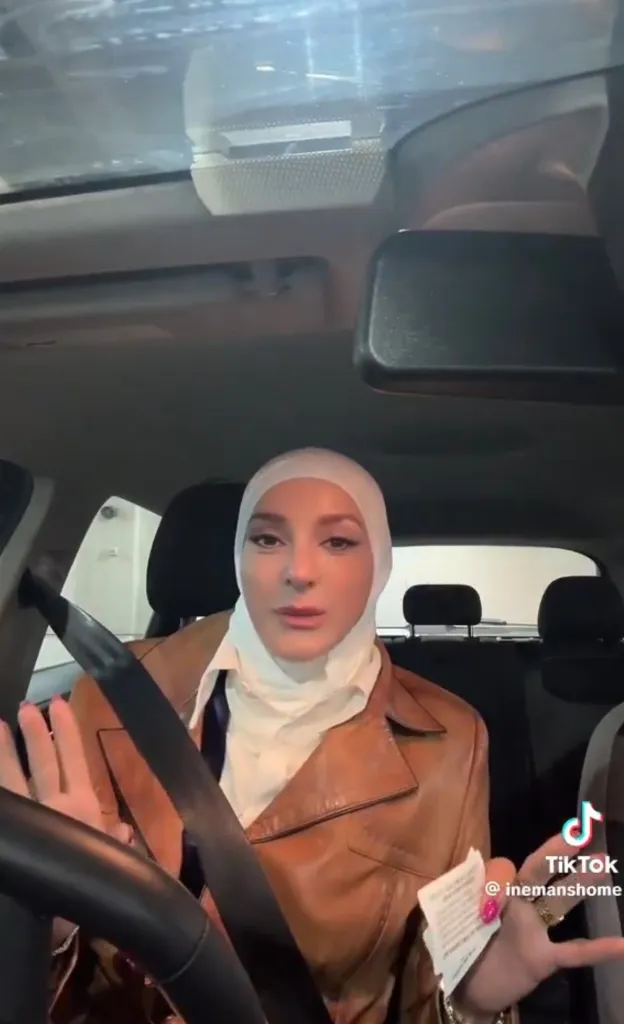 Iman Masoud, wearing a white hijab and brown jacket, in a car, speaking with her hands while holding a small piece of paper.