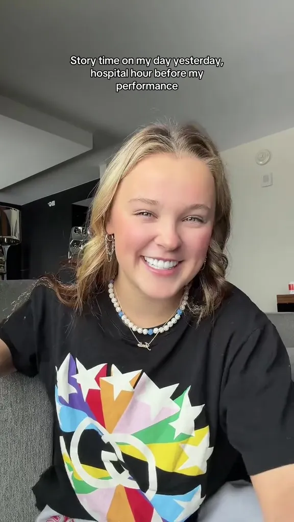 JoJo Siwa sitting and smiling, wearing a black t-shirt with a colorful star design.