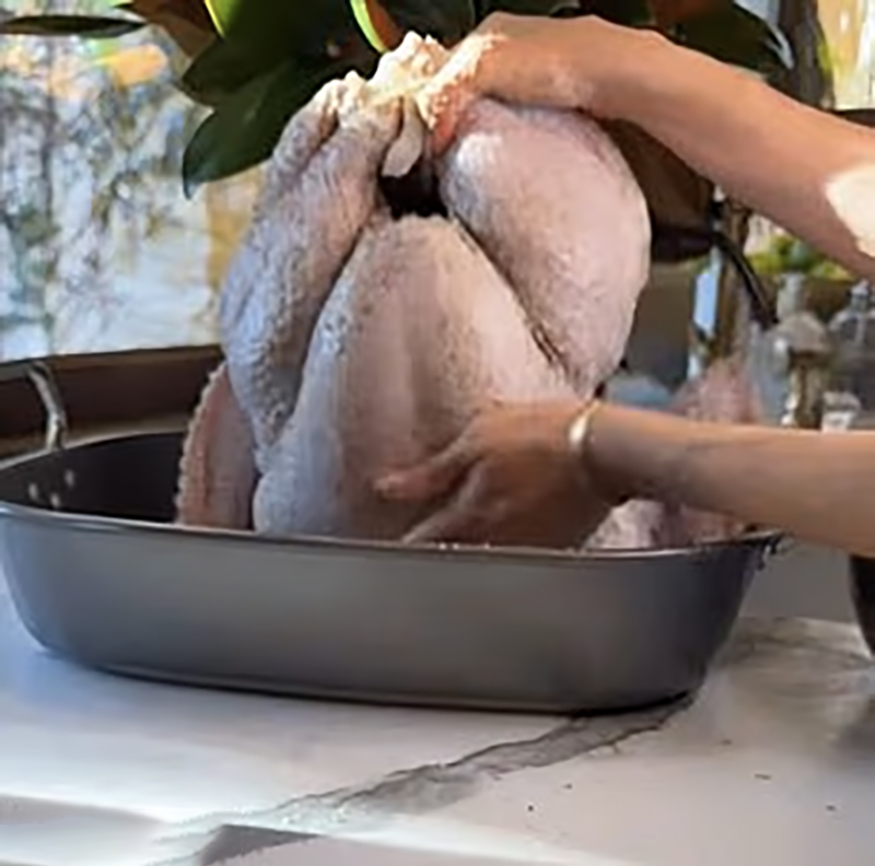 Meghan Markle's hands seasoning a raw turkey in a roasting pan.