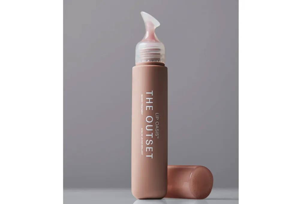 A tube of The Outset Lip Oasis Glossy Treatment lip gloss, with its applicator visible and cap placed on the surface next to it.
