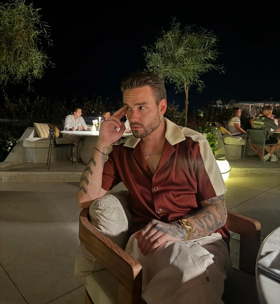 Liam Payne in a red and white collared shirt, light-colored pants, and a gold watch, sitting in an outdoor setting at night, with other people and trees in the background.