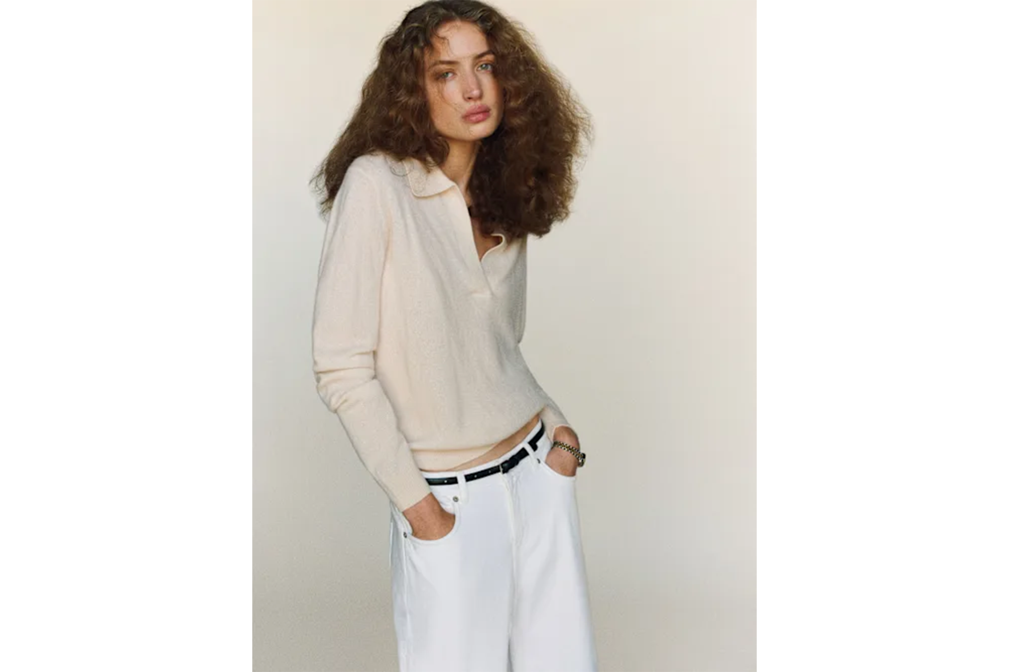 A woman with long curly hair wearing a cream-colored collared shirt, white pants, and a black belt.
