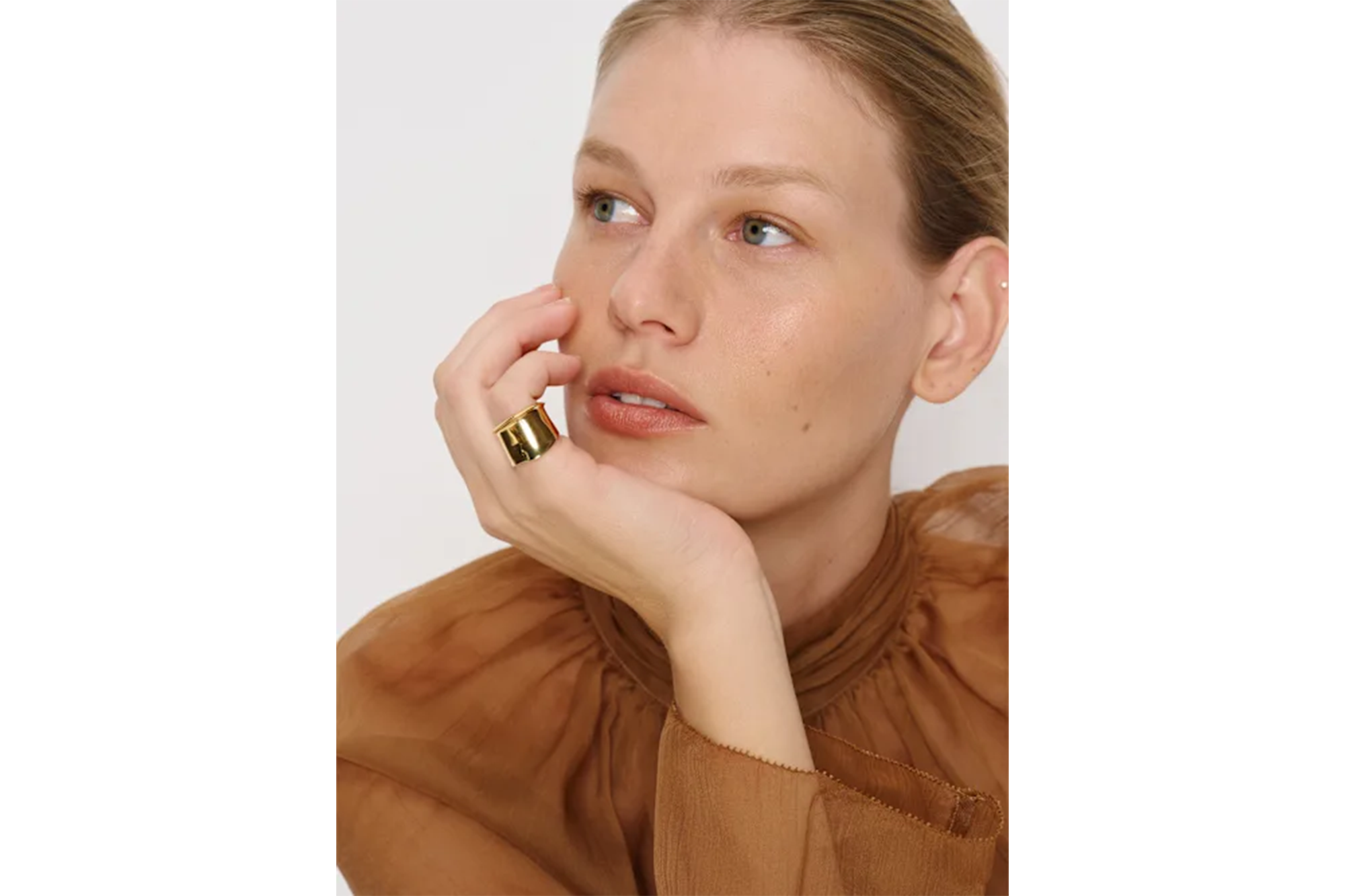 Woman with blonde hair wearing a brown blouse and a gold ring, looking to the left.