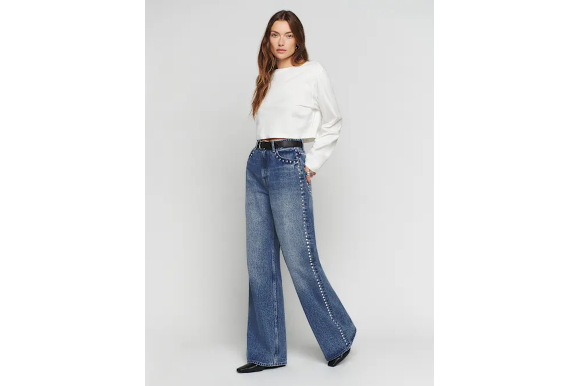 A model wearing a white long-sleeved crop top and wide-leg blue jeans with stud detailing.