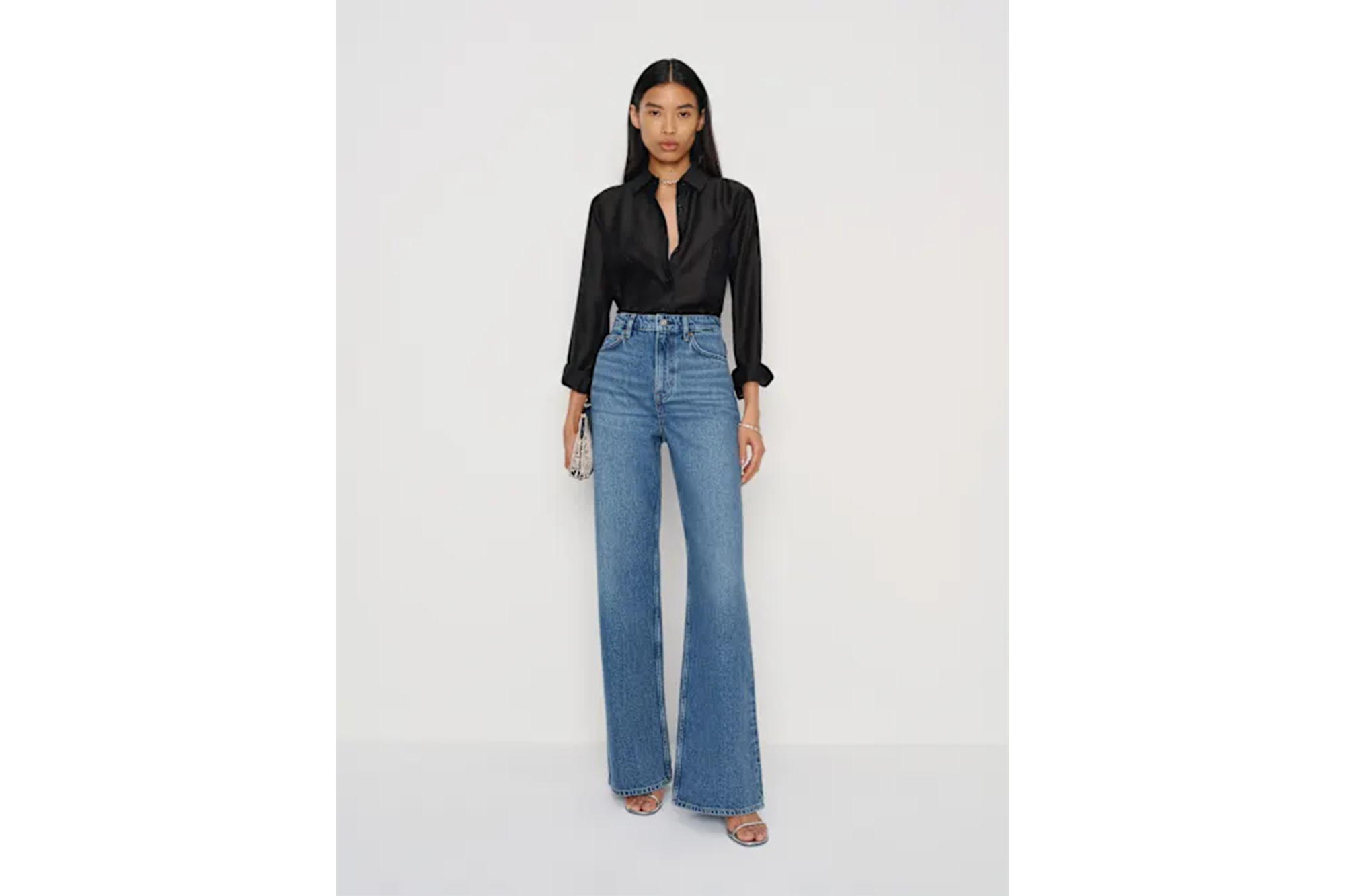 A woman in a black shirt and high-waisted wide-leg jeans, holding a small silver purse.