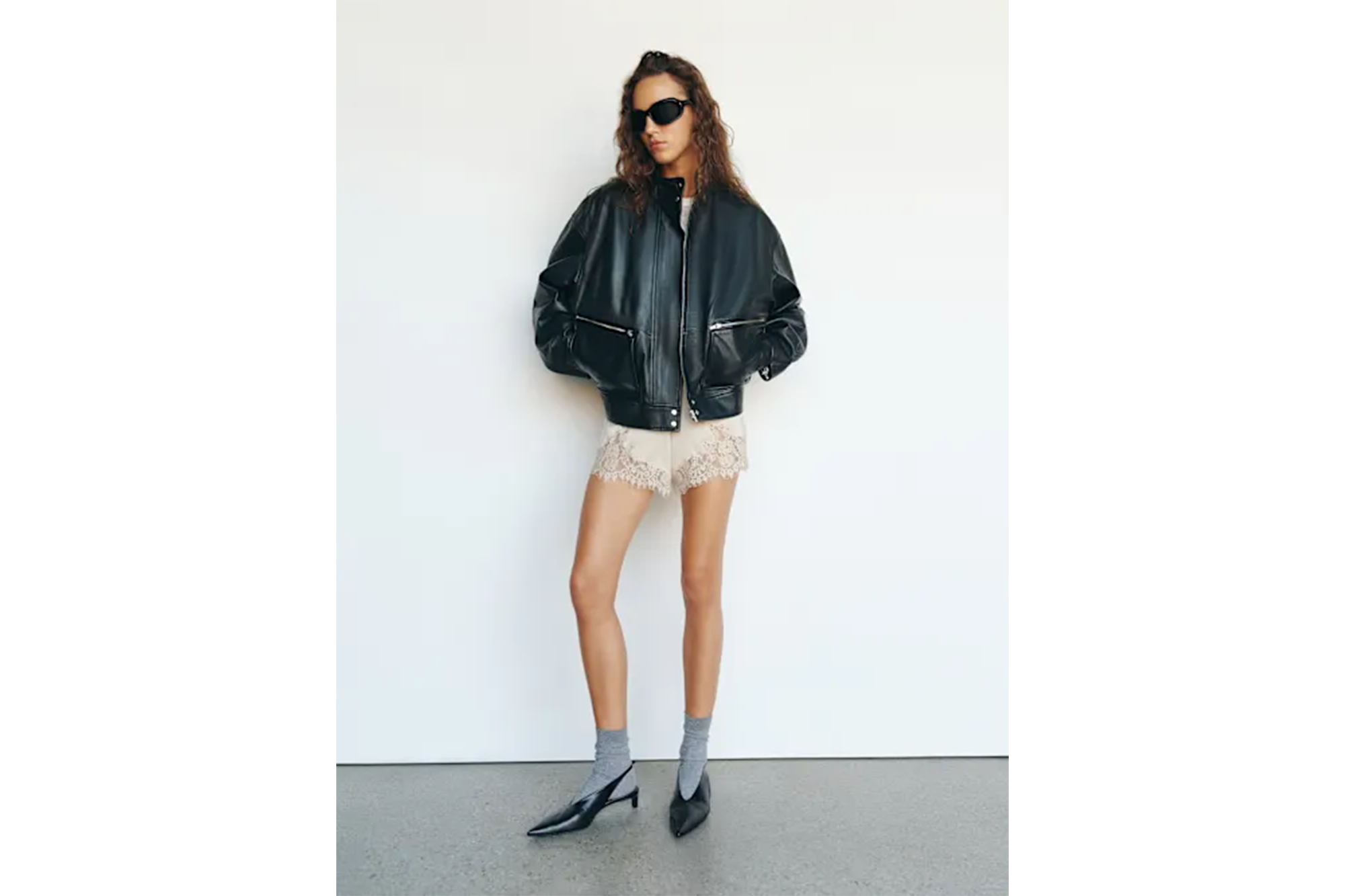 A person wearing a black bomber jacket, cream lace shorts, black kitten heels, and sunglasses.