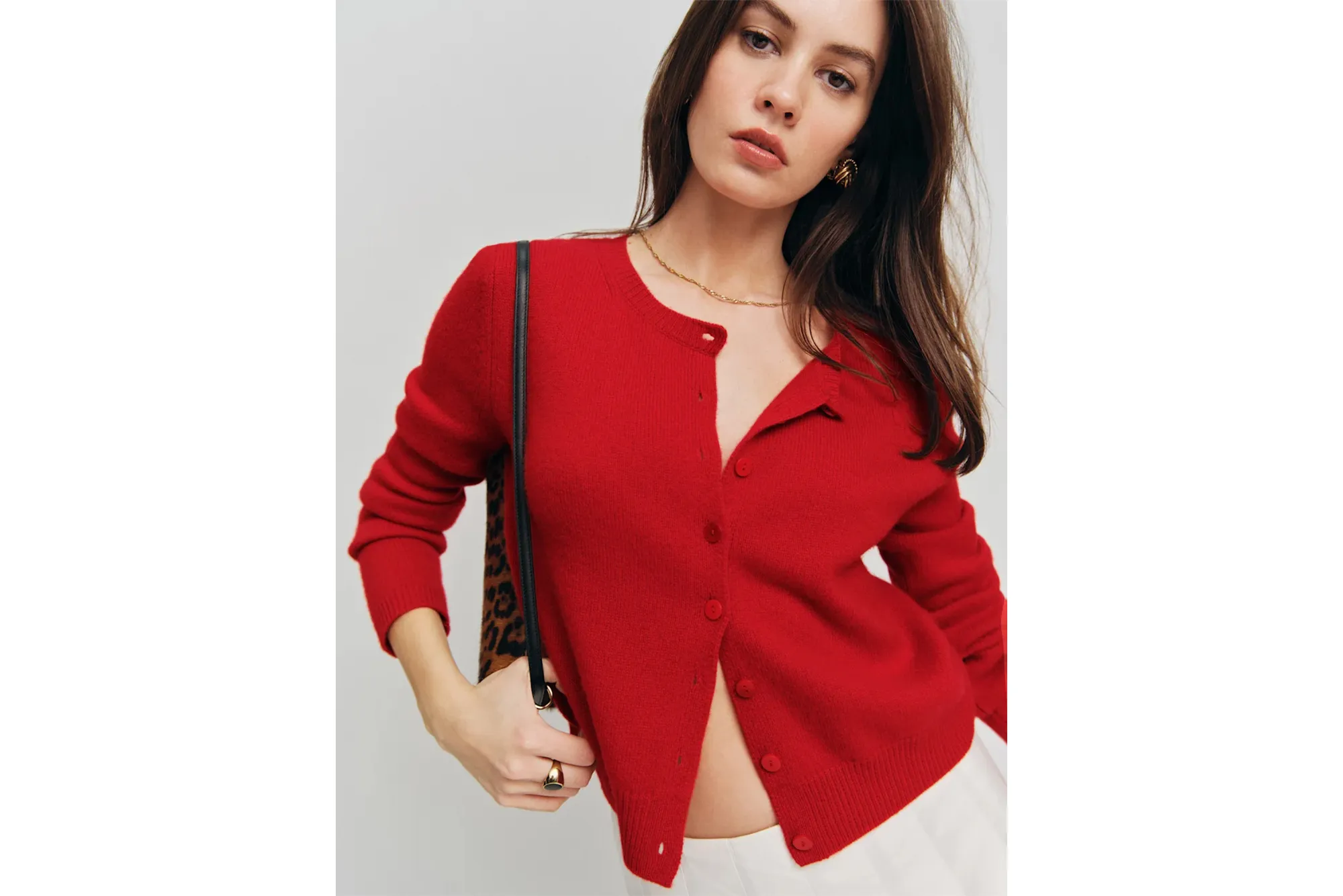 A woman wearing a red Clara Cashmere Cardigan, a gold chain necklace, gold earrings, and white pants, holding a leopard print bag.