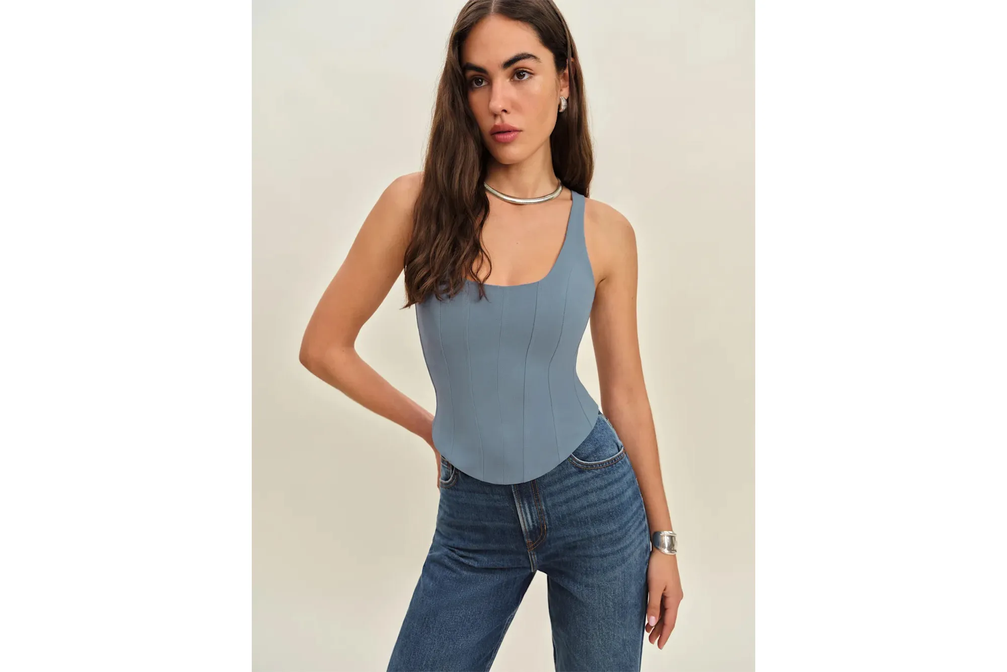 A woman models the Reformation Seren Top, a light blue bustier-style top with a square neckline, paired with blue jeans.