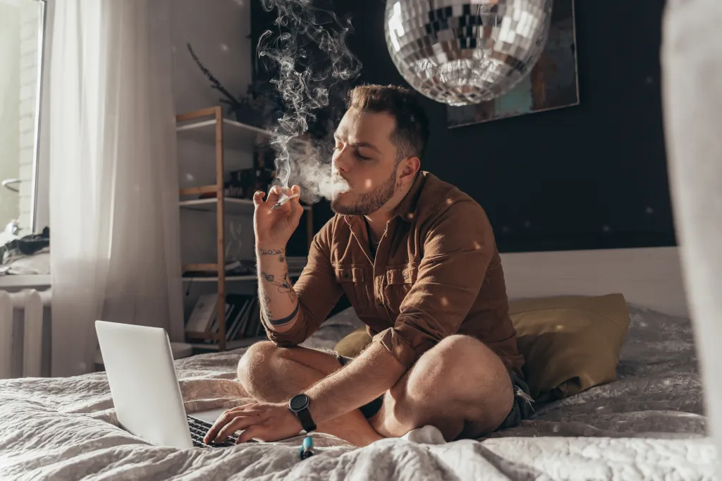 Man blowing out puffs of smoke from a hand-rolled cigarette while sitting on a bed with a laptop.