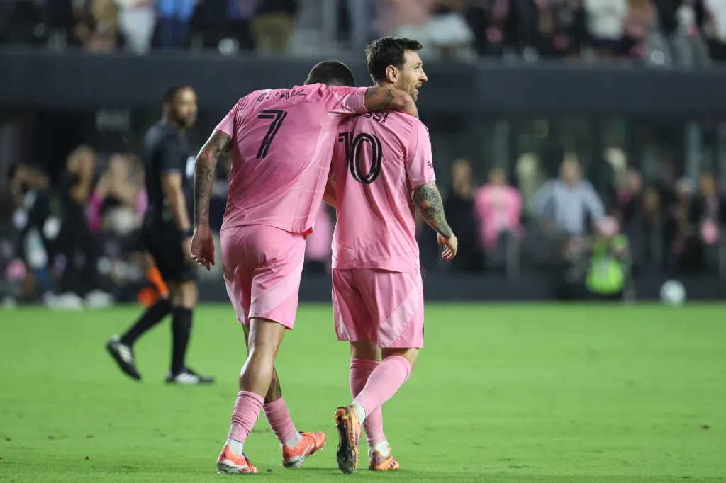 Rodrigo De Paul and Lionel Messi in pink uniforms on a soccer field.