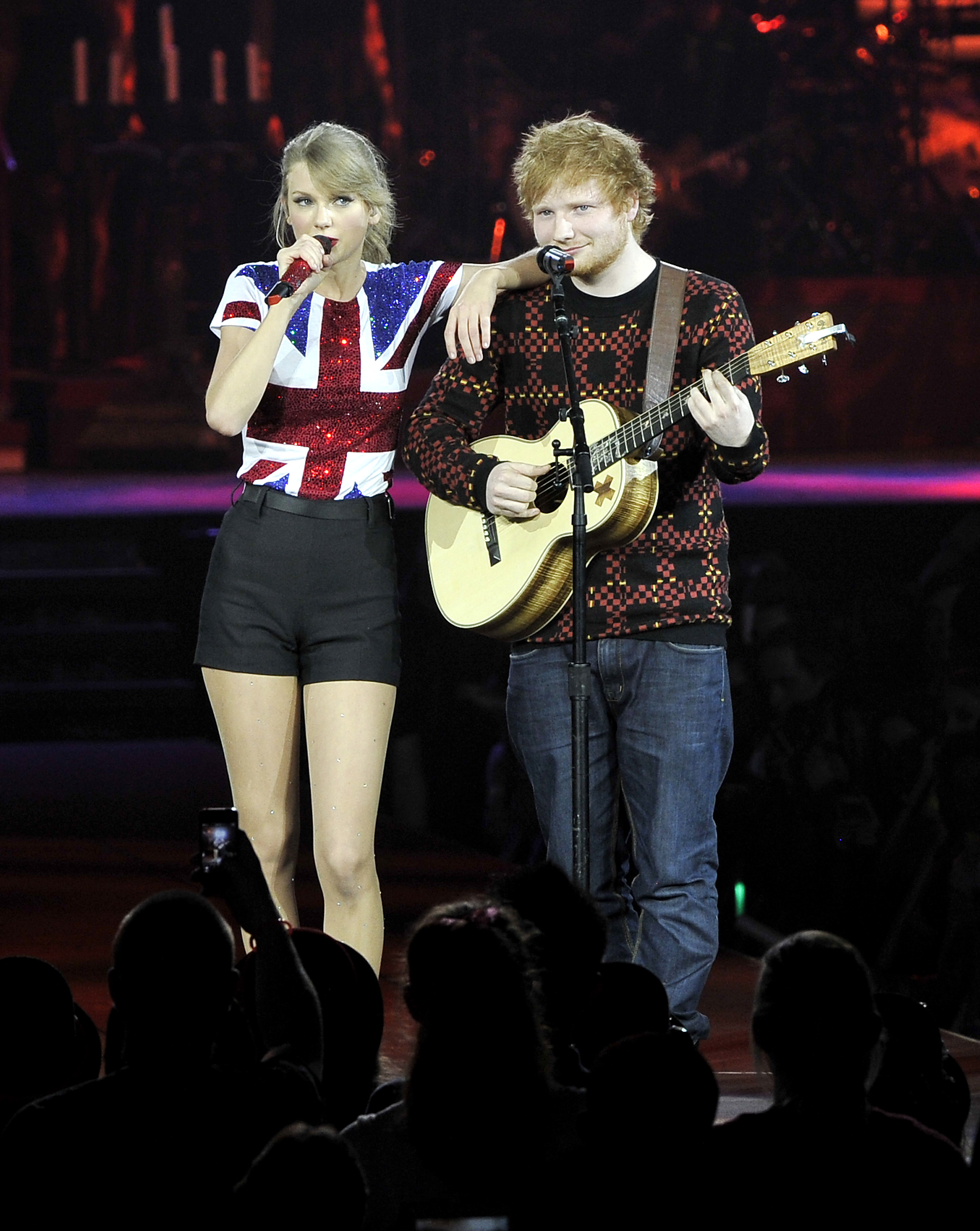 Taylor Swift in a Union Jack shirt with Ed Sheeran playing guitar on stage.