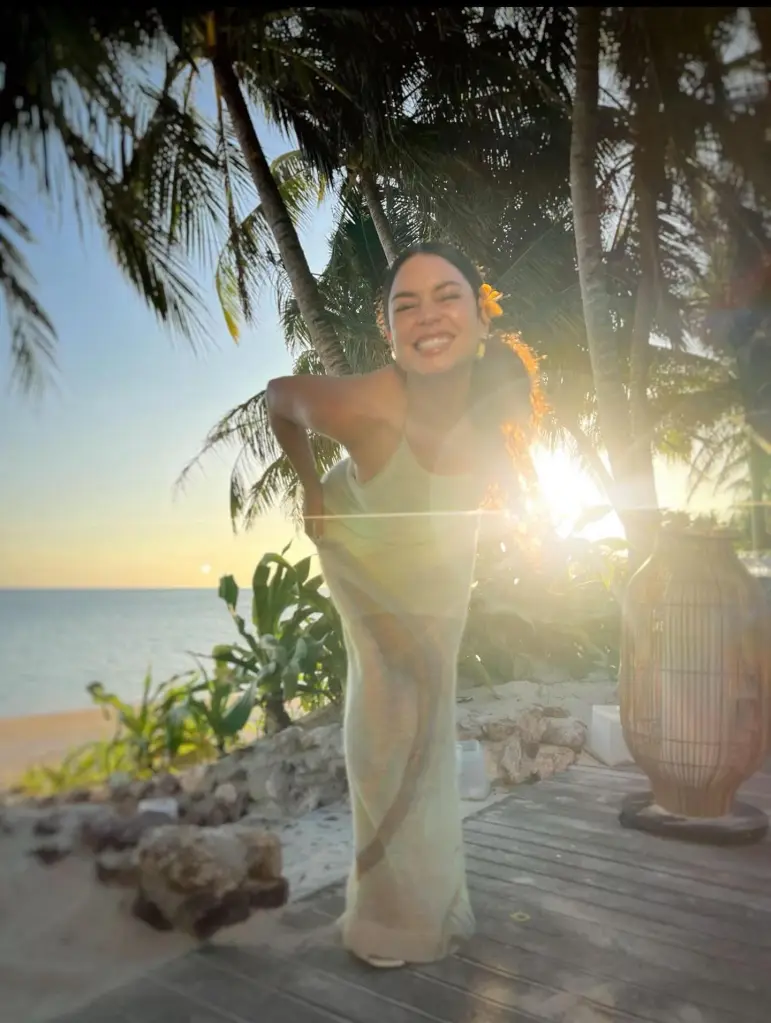 Vanessa Hudgens smiles brightly while posing on a beach with palm trees and an ocean sunset in the background.