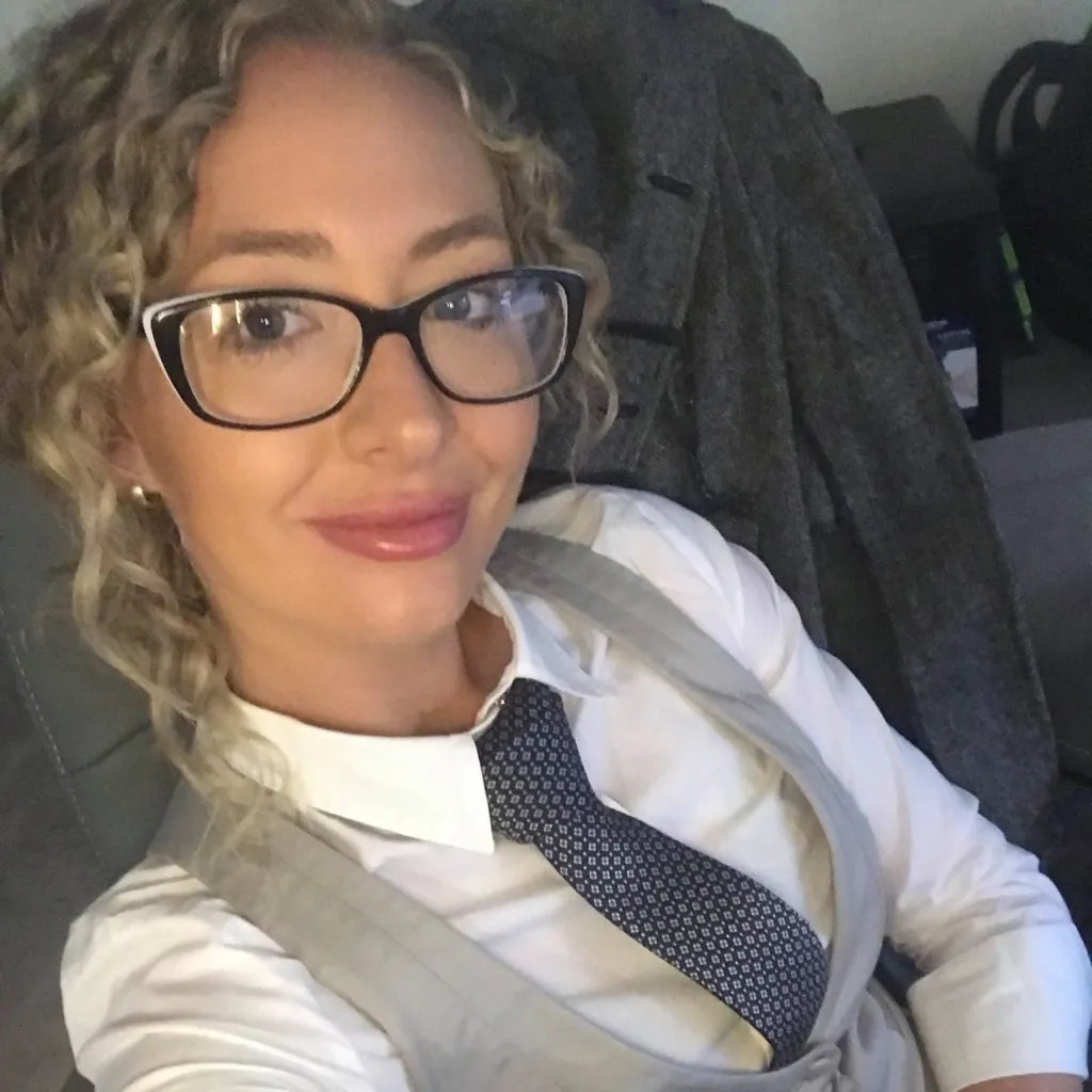 A woman with curly blonde hair and glasses wearing a white shirt, a gray vest, and a patterned blue tie.