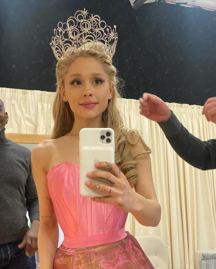 Ariana Grande as Glinda in a pink dress and silver crown taking a selfie.
