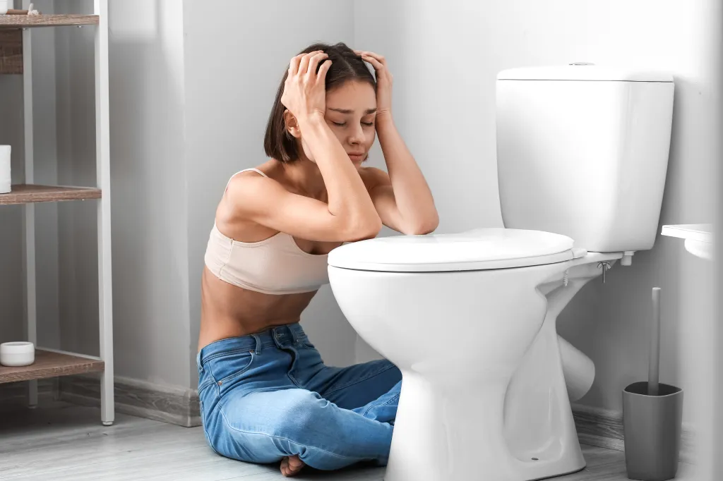 A distressed young woman, sitting on the floor next to a toilet, clutching her head in her hands.