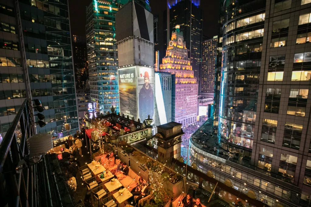The Knickerbocker St Cloud Ball View Rooftop at night, surrounded by brightly lit buildings, and featuring people dining at tables decorated with string lights.