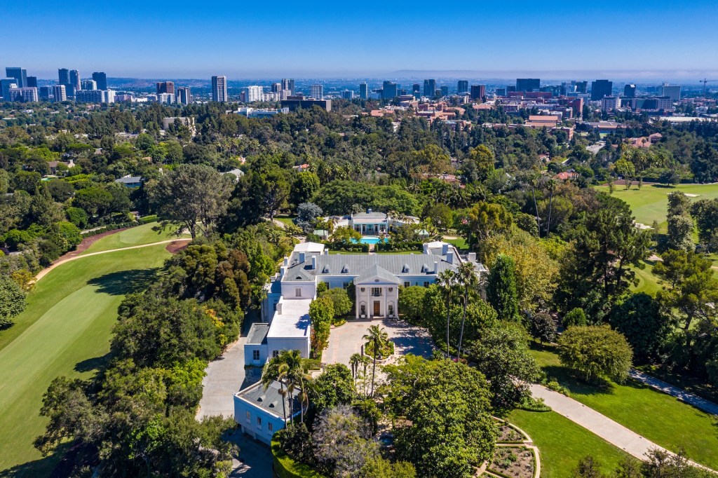 Aerial view of a large white 1930s Bel-Air estate surrounded by green trees and a city skyline in the background.