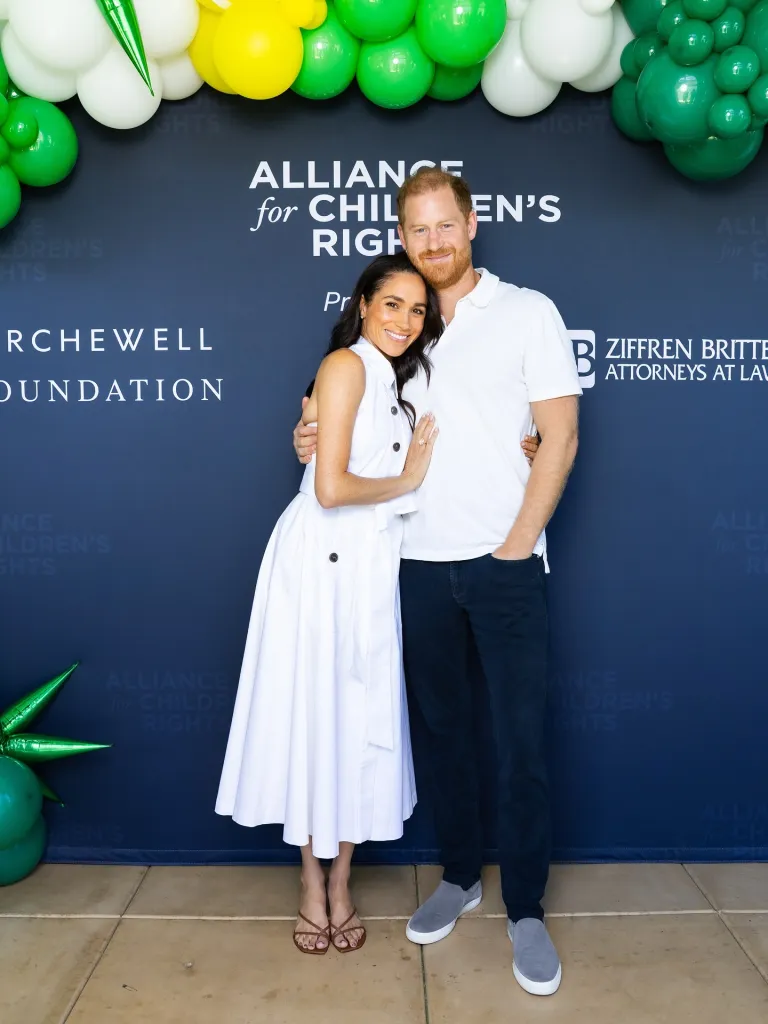 Meghan Markle and Prince Harry posing for a photo in front of a banner for the Alliance for Children's Rights and Archewell Foundation.