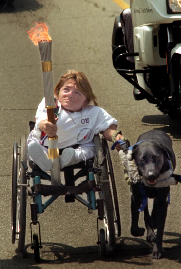 Melanie Watson, in a wheelchair, holding the Olympic torch, is pulled by her dog, Roamer.