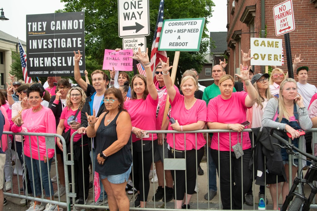 A group of people, many wearing pink shirts, holding protest signs behind a metal barrier.