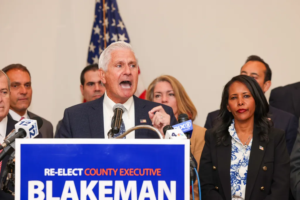 Nassau County Executive Bruce Blakeman speaking at a podium with microphones, with other people around him, and an American flag in the background.