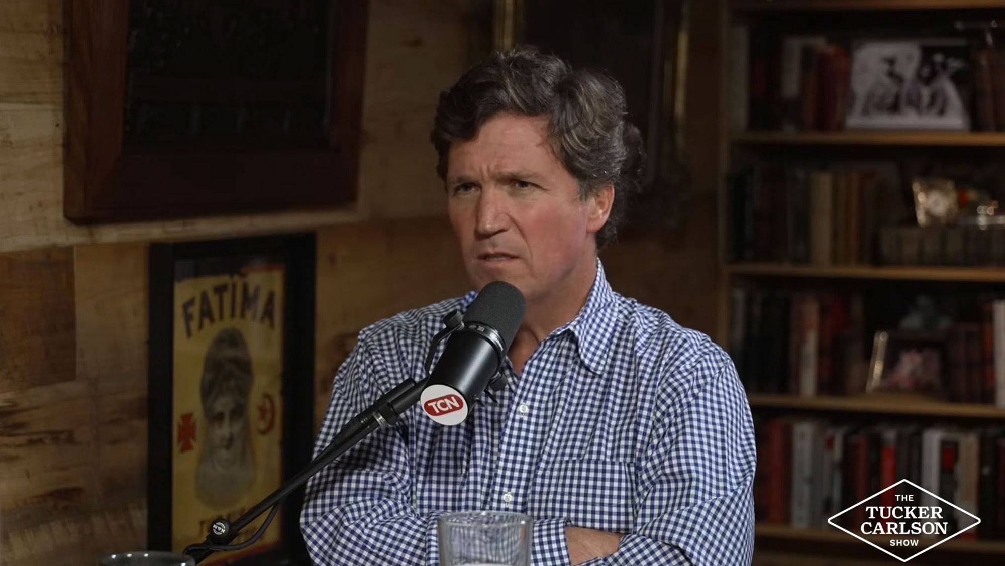 Tucker Carlson in a blue gingham shirt, speaking into a microphone.