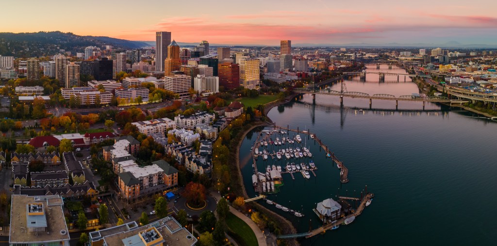 Aerial panoramic view of downtown Portland, Oregon at sunset, featuring the Willamette River, bridges, and city skyline.