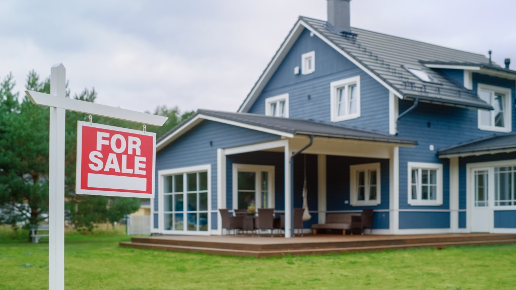 A red For Sale sign in the foreground of a large blue house with a porch and yard.